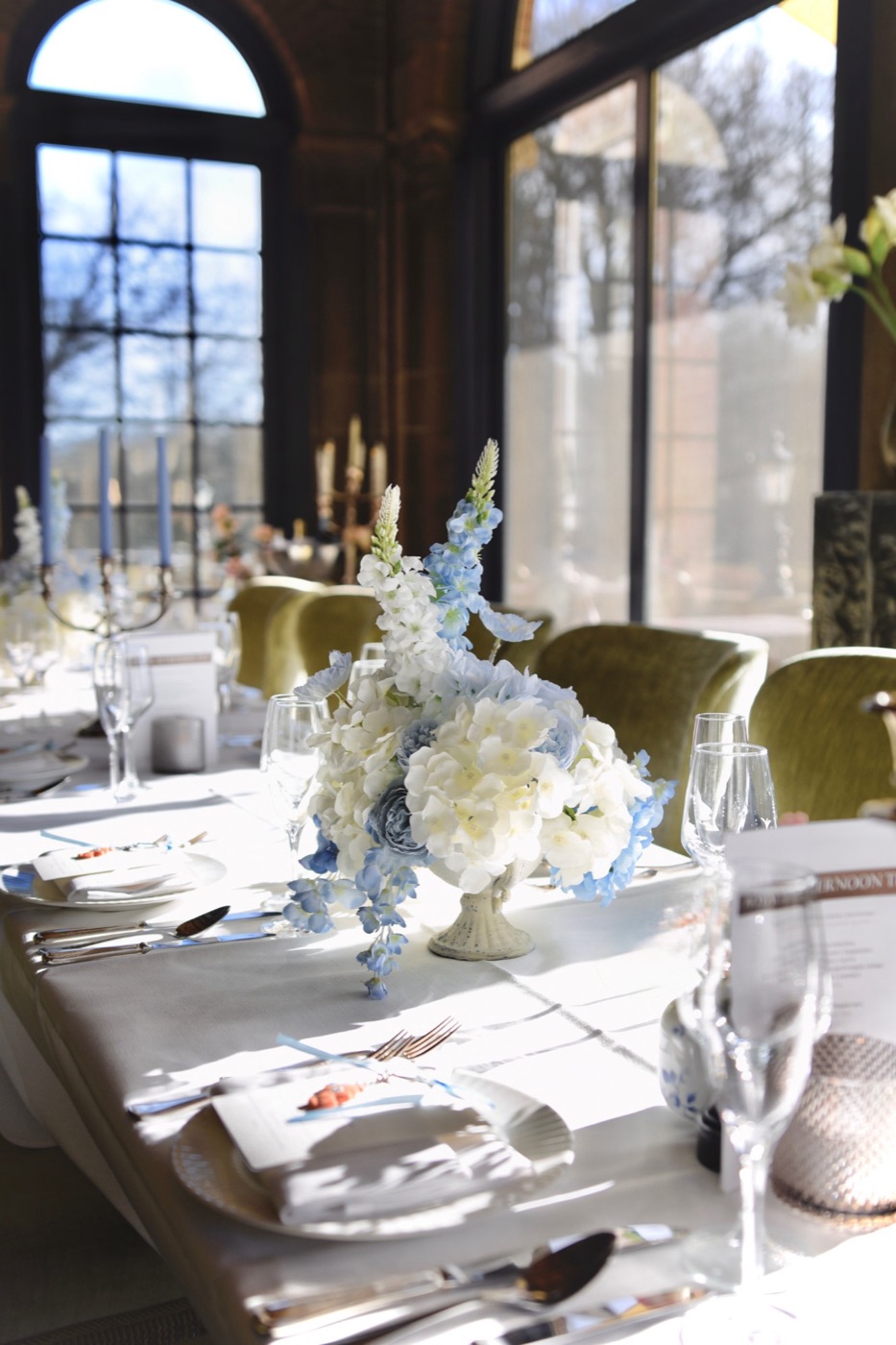 Blauw-wit bloemstuk met delphinium op een gedekte tafel in zonlicht voor hoge ramen