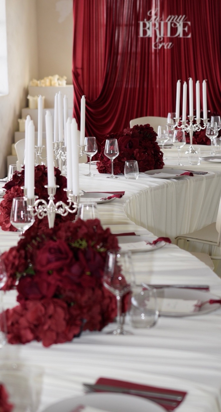 Lange feesttafel met bordeauxrode bloemstukken, kandelaars en gedrapeerd tafelkleed