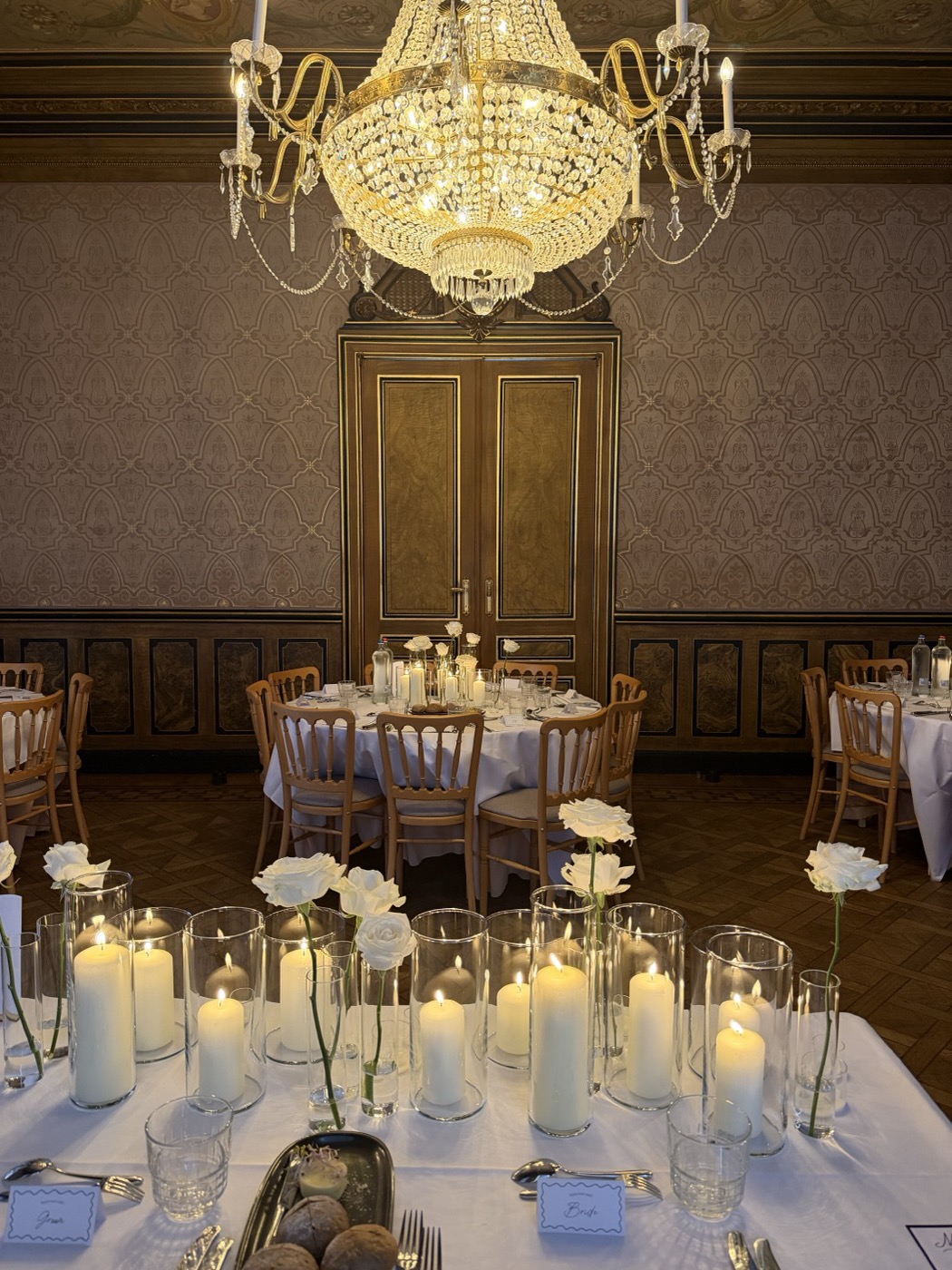 Tafels met kaarsen in glazen cilinders en losse witte rozen, met een kroonluchter in een klassieke zaal