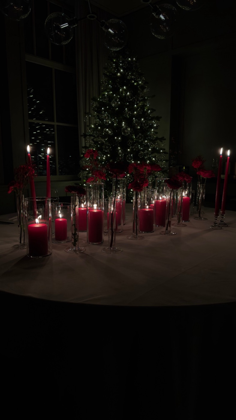 Ronde tafel met rode kaarsen in glazen, rode bloemen en een verlichte kerstboom op de achtergrond
