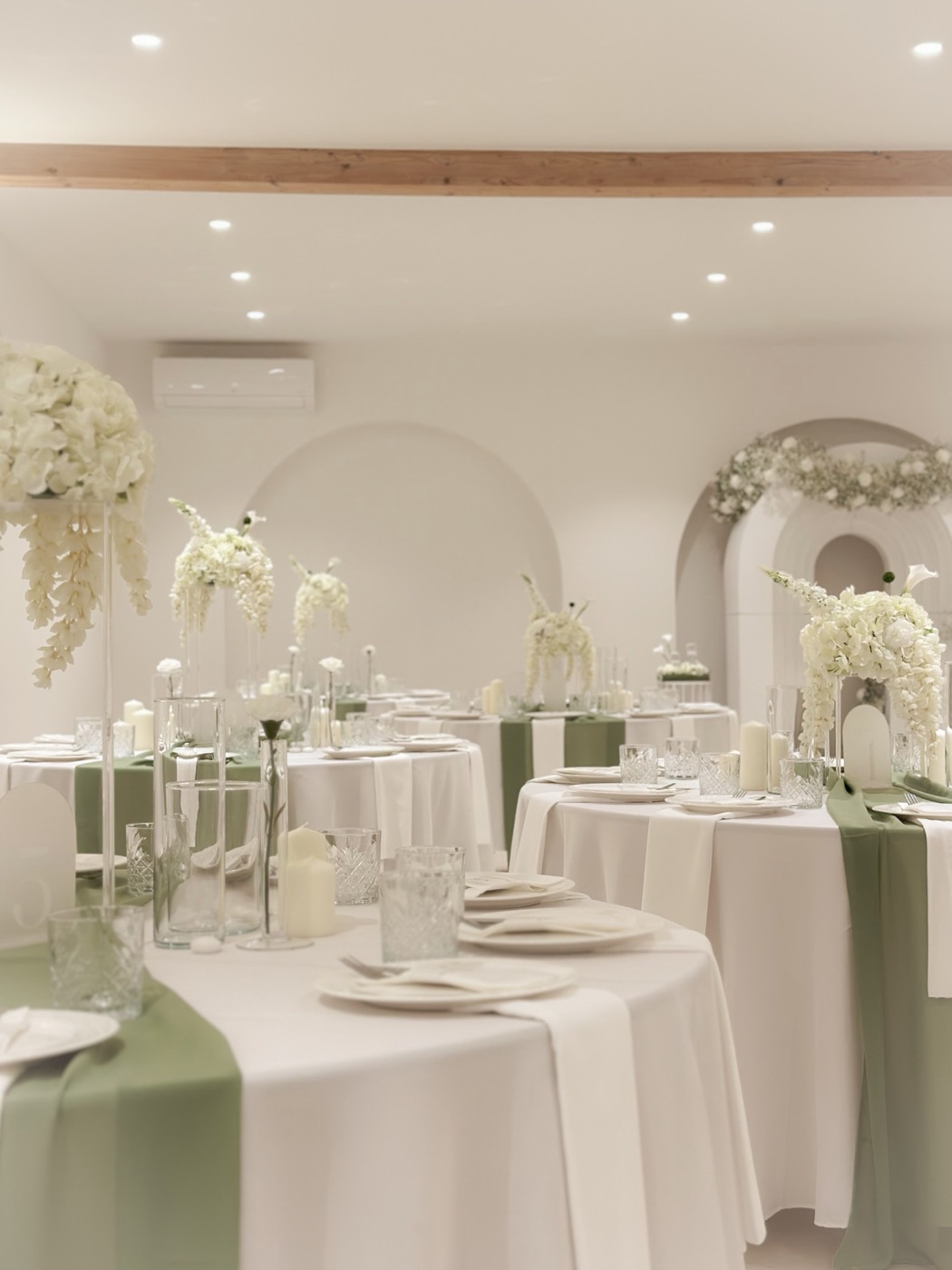 Ronde tafels met witte tafelkleden, saliegroene lopers en hoge witte bloemstukken in een lichte zaal
